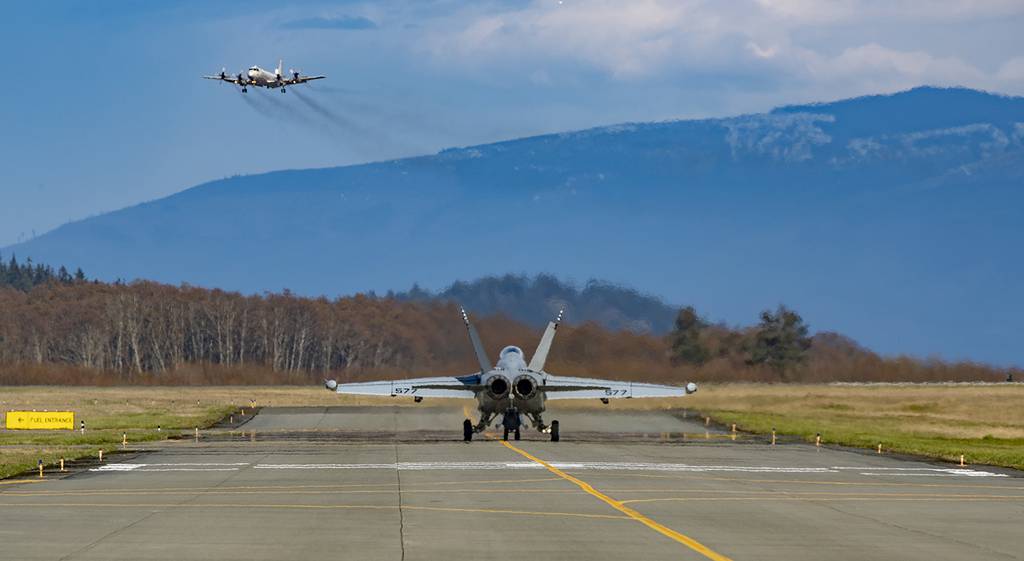 An EA-18G Growler taxis toward the runway on March 29, 2019, at Ault Field at Whidbey Island, Wash., as a P3C Orion performs a low approach.